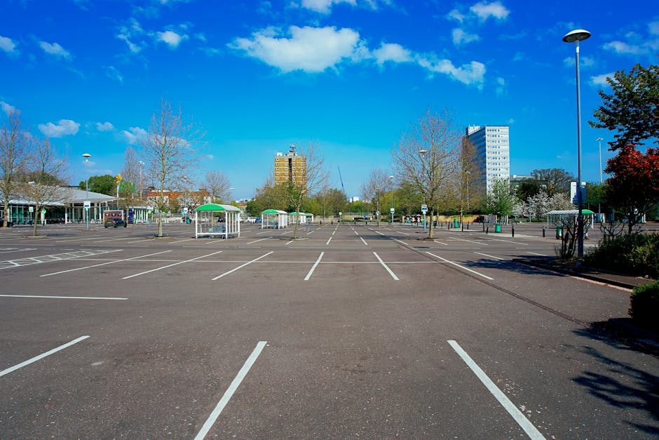 A large, mostly empty outdoor parking lot with clearly marked white parking spaces on dark asphalt. In the background, there are a few small green-roofed shelters and a few parked cars. Surrounding the parking area are leafless trees, some with sparse blossoms, and a mix of modern high-rise buildings and smaller structures. Tall streetlights are positioned along the edges of the lot, with one prominently visible on the right side. The sky above is bright blue with scattered white clouds, indicating clear weather and good natural light. The scene is neutral and professional, offering a clean environment that could be associated with alternative waste handling or private collection sites, relevant to rubbish removal services like those of Rubbish Clearance Watford. The setting appears to be in a city or town environment, with no visible signage or specific objects indicating ongoing activity, emphasizing the spaciousness suitable for outdoor waste storage or clearance operations.