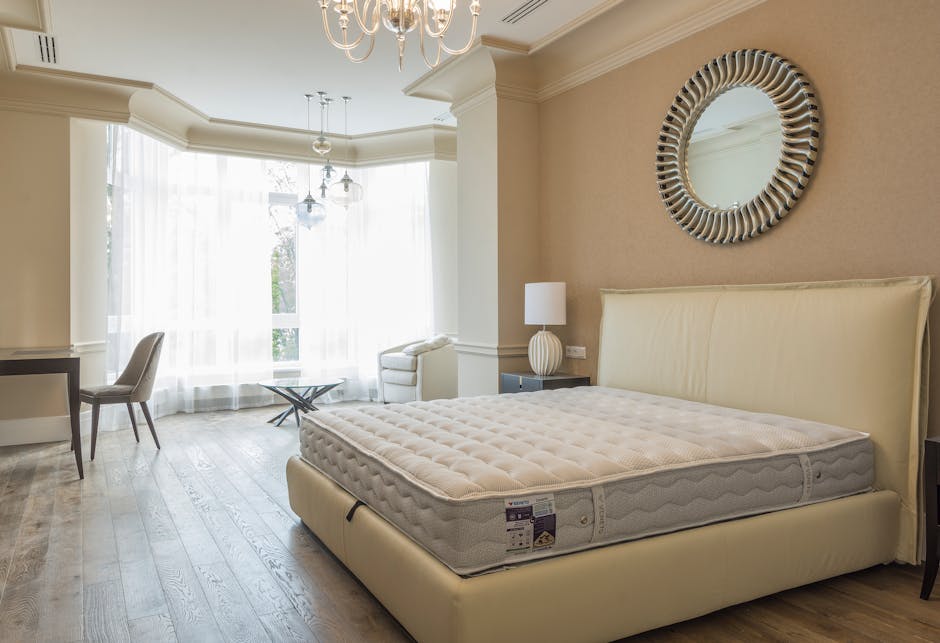 An interior bedroom scene featuring a cream-colored upholstered bed with a visibly clean, padded headboard and a new, unused mattress with a textured surface. The bed is positioned against a beige wall with a decorative round mirror framed by a striped, metallic finish hanging above it. To the right of the bed, there is a small dark nightstand holding a white table lamp with a globe-shaped base and a white lampshade. On the left side of the room, a window with sheer white curtains allows natural light to fill the space, illuminating the light wooden flooring that extends across the room. Near the window, a white armchair and a small side table are visible, adding to the room's elegant and modern decor. The ceiling is adorned with a classic chandelier and pendant lights, and overall, the room presents a neat and well-maintained appearance suitable for residential living, shared here to illustrate a typical space where furniture removal services might be coordinated through companies like Rubbish Clearance Watford, especially when clearing out unwanted items before renovation or disposal.