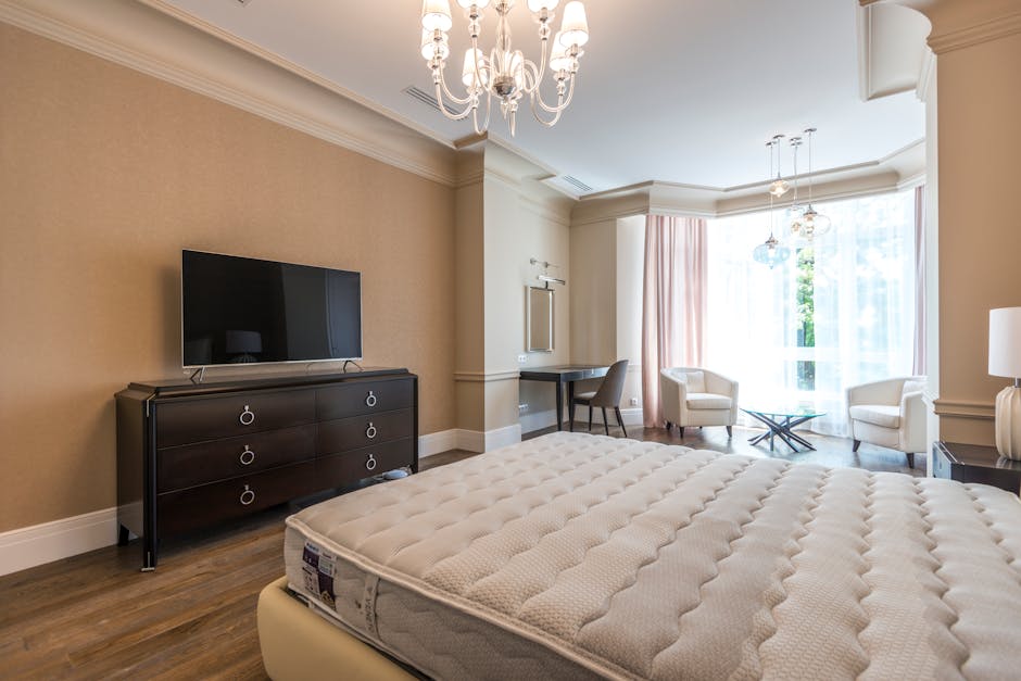 An interior bedroom scene featuring a cream-colored upholstered bed with a visibly clean, padded headboard and a new, unused mattress with a textured surface. The bed is positioned against a beige wall with a decorative round mirror framed by a striped, metallic finish hanging above it. To the right of the bed, there is a small dark nightstand holding a white table lamp with a globe-shaped base and a white lampshade. On the left side of the room, a window with sheer white curtains allows natural light to fill the space, illuminating the light wooden flooring that extends across the room. Near the window, a white armchair and a small side table are visible, adding to the room's elegant and modern decor. The ceiling is adorned with a classic chandelier and pendant lights, and overall, the room presents a neat and well-maintained appearance suitable for residential living, shared here to illustrate a typical space where furniture removal services might be coordinated through companies like Rubbish Clearance Watford, especially when clearing out unwanted items before renovation or disposal.