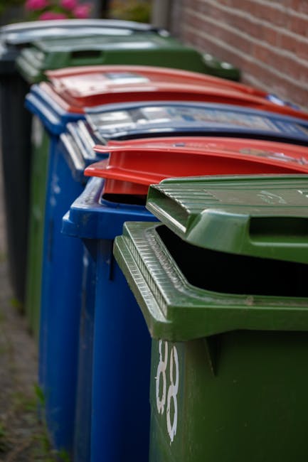 A row of five large wheelie bins positioned along a paved pathway beside a brick wall, with each bin having a different colored lid: green, blue, red, dark blue, and another green. The green bin in the foreground has a slightly raised lid, revealing a textured surface within. The blue, red, and dark blue bins are lined up behind, with their lids closed, showing smooth finishes and uniform coloring. The bins are situated outdoors, with an environment suggesting an urban or residential setting, likely for waste collection or rubbish disposal. The scene is illuminated by natural light, capturing the distinct colors and textures of the plastic bins, commonly used for private waste management such as rubbish clearance services, aligning with the context of independent waste collection or alternatives to council rubbish collection.