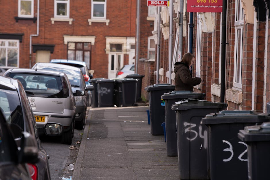 A street scene in a residential area with a row of dark grey wheelie bins placed along the sidewalk in front of red-brick houses with white window frames and decorative stone lintels. A woman, dressed in a brown coat, is standing next to one of the bins, possibly organizing or disposing of rubbish. Several vehicles are parked along the curb, including a silver hatchback in the foreground and additional cars further down the street. The pavement has some litter scattered about, and the houses behind the bins feature mixed signage and window displays, indicating commercial or mixed-use properties. The lighting suggests a cloudy day with soft natural light illuminating the scene. The presence of multiple bins and parked cars emphasizes an urban setting where private rubbish collection services, such as those offered by Rubbish Clearance Watford, manage waste disposal outside the scope of local council kerbside collections. The scene illustrates a typical site for on-site rubbish handling and private waste clearance in a residential neighbourhood.